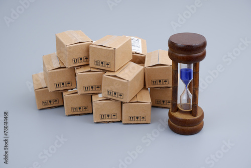 Delivery time concept. Hourglass and many carton boxes on gray background.	