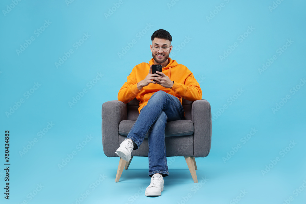 Happy young man using smartphone on armchair against light blue background Happy young man using smartphone on armchair against light blue background