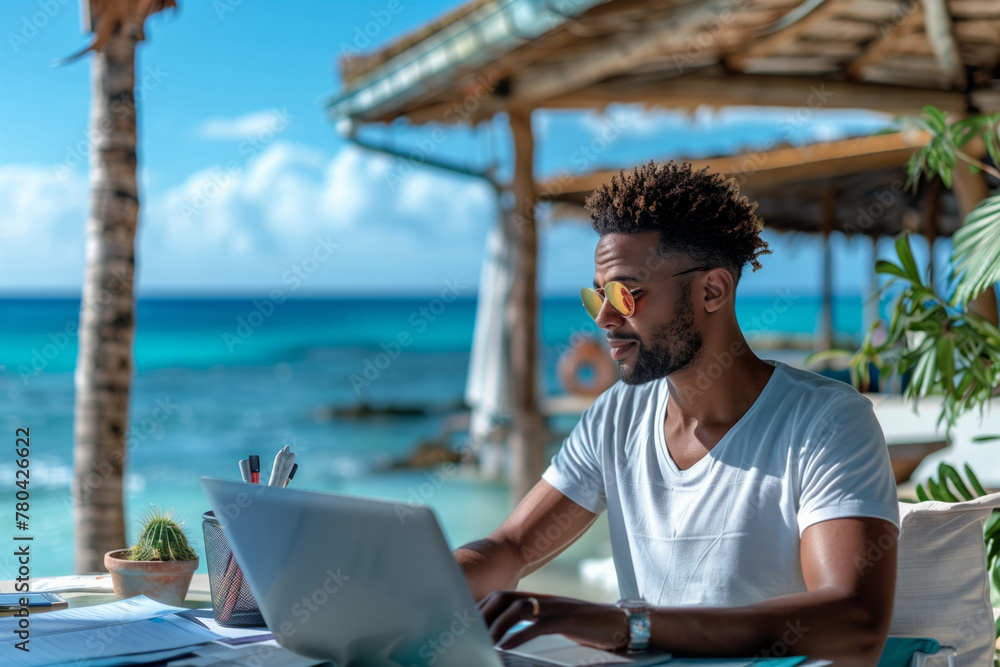 A dark skin young man in a T-shirt works on a laptop at a table on the beach in the summer.
