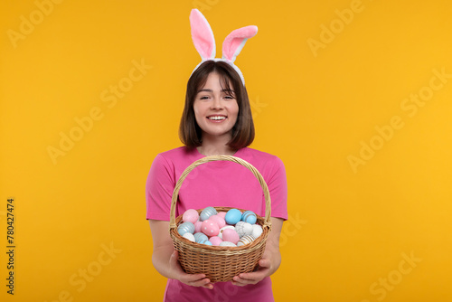 Easter celebration. Happy woman with bunny ears and wicker basket full of painted eggs on orange background