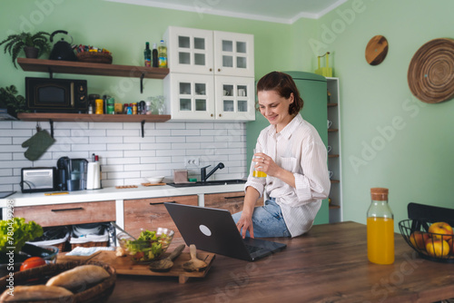 Young cheerful woman using laptop at home in kitchen, working from home