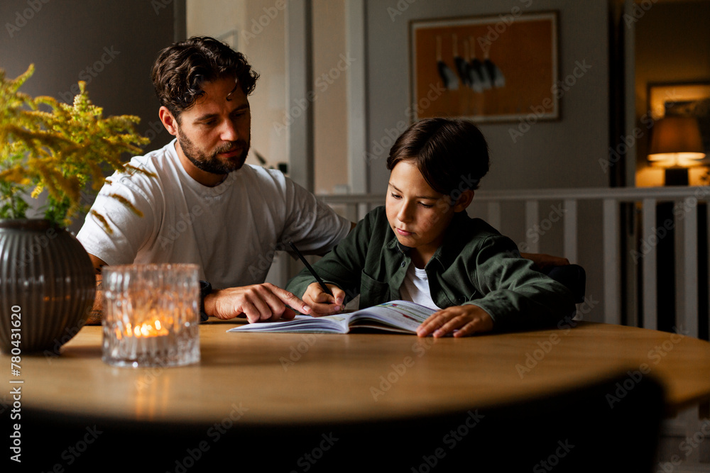 Man assisting son writing in book while doing homework at table Man assisting son writing in book while doing homework at table