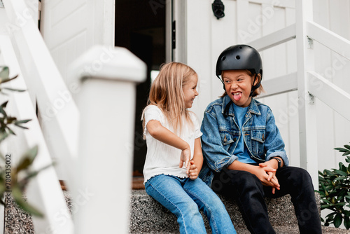 Boy wearing cycling helmet sticking out tongue and making face in front of sister sitting at front stoop photo