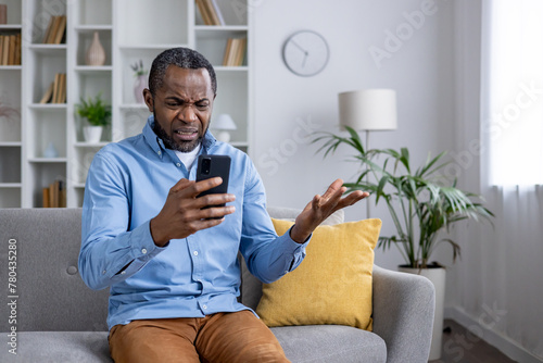 Perplexed African American man sitting in a living room, holding a smartphone and gesturing in confusion over a deceptive message. © Liubomir
