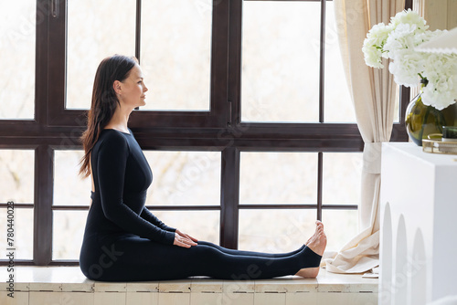 Woman performs Dandasana exercise at home, staff pose, practices yoga in sportswear sitting on windowsill, full-length view