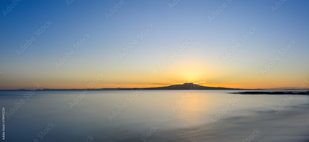 Golden glow at the crest of Rangitoto Island as the sun is rising above the horizon. Milford Beach. Auckland.