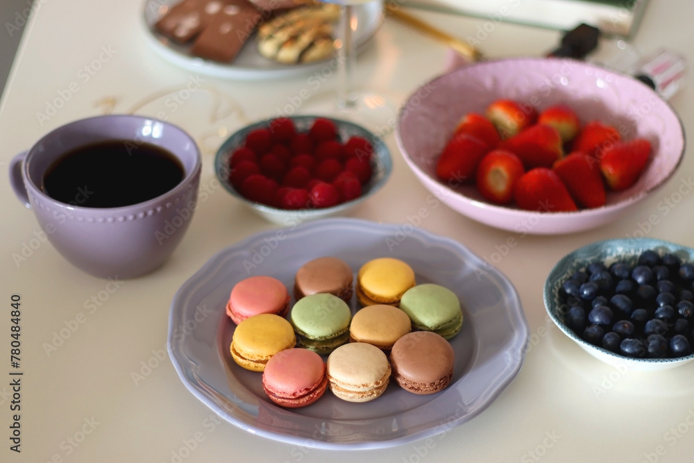 Plate of pastel macarons, cookies and chocolate, cup of tea of coffee, glass of bubble water, various berries, books and accessories on the table. Selective focus, pastel colors. Plate of pastel macarons, cookies and chocolate, cup of tea of coffee, glass of bubble water, various berries, books and accessories on the table. Selective focus, pastel colors.