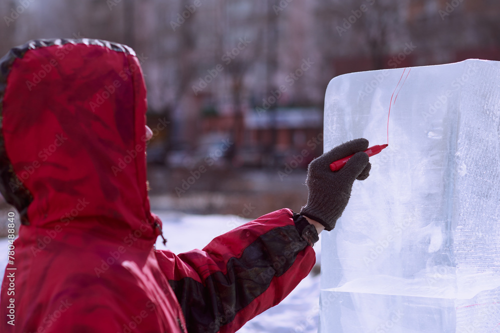 A sculptor draws a red line on an ice block. Preparing to create a sculpture at the city winter festival of artistic ice figures. - obrazy, fototapety, plakaty 