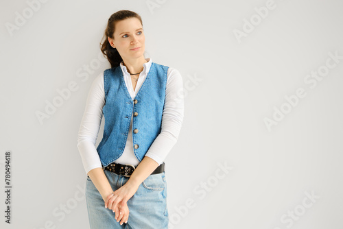 Relaxed woman in denim vest, jeans and cotton shirt leaning against white wall