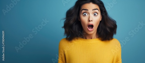 Shocked young brunette in yellow sweater against blue wall reacts to surprising news, holding her breath.