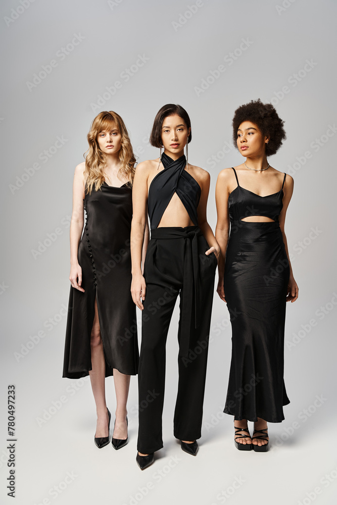 Obraz premium Three women of diverse backgrounds stand next to each other in elegant black dresses on a grey studio background.