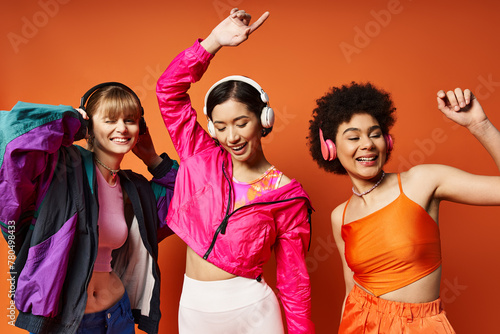 Three diverse women in headphones smiling for a vibrant orange studio photo. © LIGHTFIELD STUDIOS