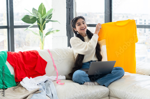 Smiling young indian woman sitting on a cozy couch, holding yellow t-shirt and using laptop to shop online, looking cheerful and delighted with her purchase.
