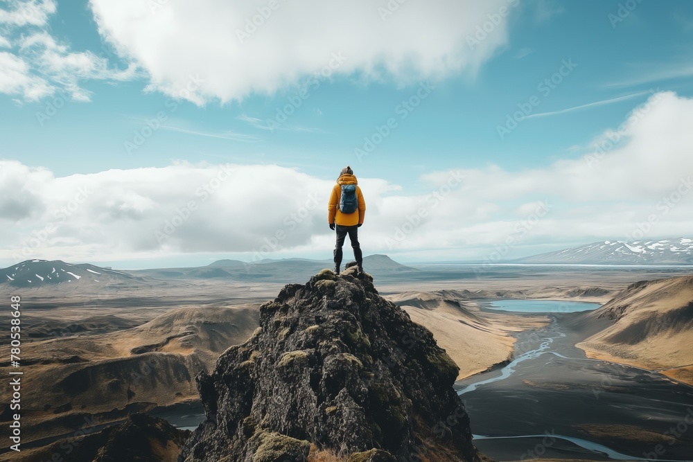 A person is standing on a mountain top, looking out over a vast landscape. The sky is clear and blue, and there are no clouds in sight. The person is wearing a yellow jacket and has a backpack on