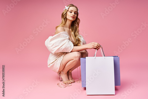A young, blonde woman gracefully kneels down with a shopping bag in a studio setting.