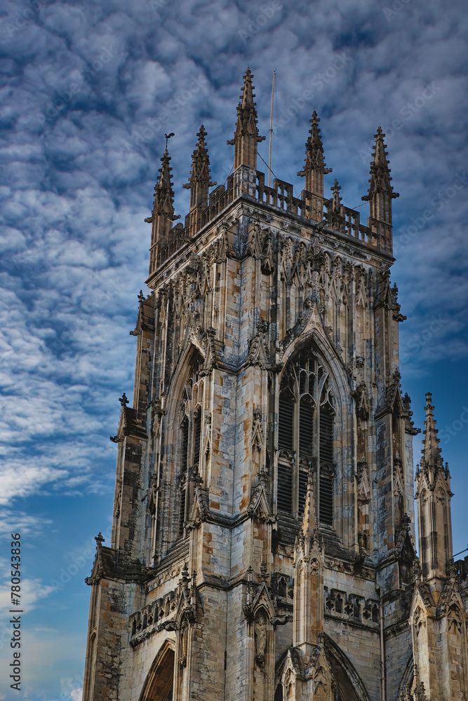 Obraz premium Gothic cathedral tower against a dramatic cloudy sky, showcasing intricate architectural details and spires, ideal for historical or religious themes in York, North Yorkshire, England.
