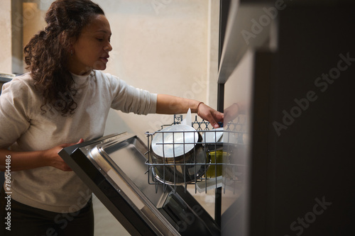 Charming multi ethnic woman loading, unloading, emptying out, putting dishes inside baskets in automatic dishwasher at home kitchen photo