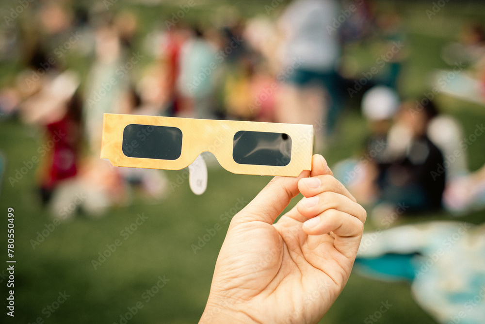 Fototapeta premium Hand holding paper solar eclipse with blurry crowd people watching totality show picnic yard, Dallas, Texas, April 8, scratch resistant polymer lenses filter out harmful ultraviolet, infrared ray