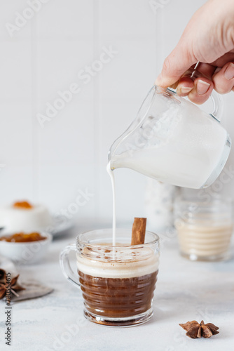 A woman's hand pours milk from a milk jug into a glass mug with black coffee, light concrete background. Kinfolk style composition.