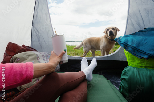 An unrecognizable woman in a tent drinks tea from a mug overlooking the lake. The dog is standing next to the human