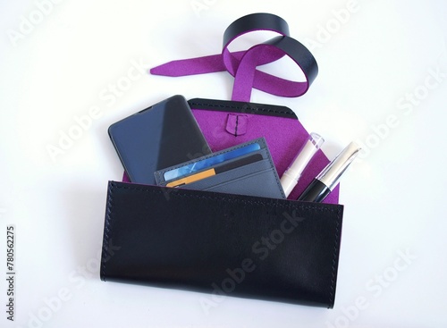 Open female leather clutch bag with purple interior on the strap. Smartphone, cardholder, lipstick and mascara looks out of small black handbag. White background, flat lay, top view, space for text,