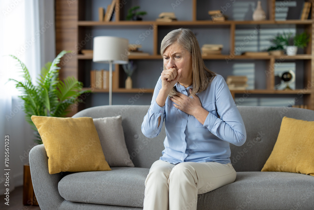 Senior stylish woman feeling sick and covering mouth with clenched fist for stopping cough while sitting on sofa with cushions. Unhealthy female feeling pain in chest and suffering from flu symptoms. Senior stylish woman feeling sick and covering mouth with clenched fist for stopping cough while sitting on sofa with cushions. Unhealthy female feeling pain in chest and suffering from flu symptoms.