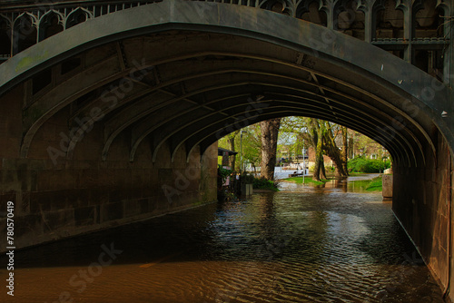 Scenic view under an old stone bridge with intricate arches, reflecting on a tranquil water surface, surrounded by lush greenery in a serene park setting in York, North Yorkshire, England.
