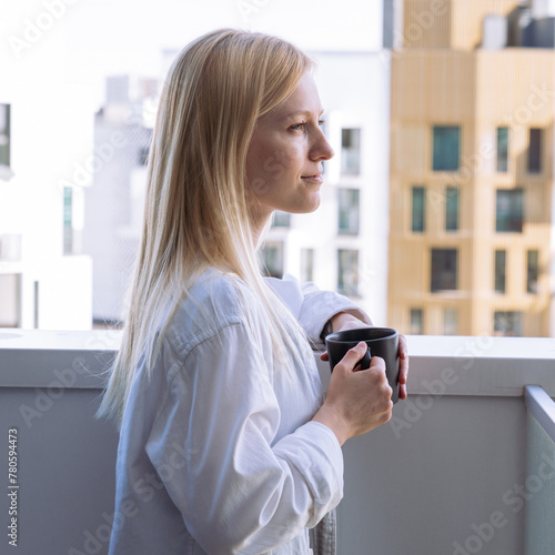 A young smiling woman drinks coffee on a balcony overlooking the new urban areas.