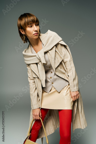 A chic young woman with short hair, donning tights and a coat, holds a fashionable handbag in a studio setting. photo