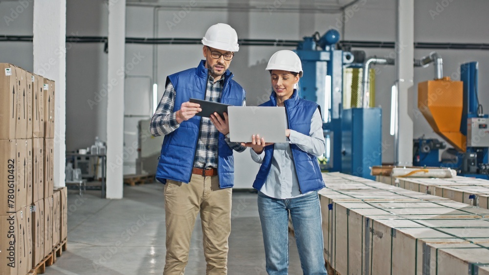 Two Caucasian workers wearing uniforms and helmets while standing in large storage or industrial facility. People actively talking together about distributing production. Using technology devices. Two Caucasian workers wearing uniforms and helmets while standing in large storage or industrial facility. People actively talking together about distributing production. Using technology devices.