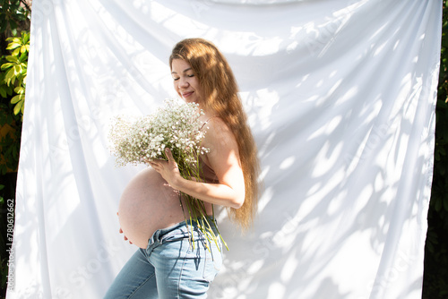 Happy Caucasian pregnant woman with long hair holding flower bouquet. Baby's breath flowers. 37 weeks pregnant woman outdoors with bare belly wearing jeans photo