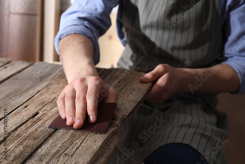 Man polishing wooden table with sandpaper indoors, closeup photo