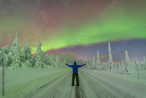 Rear view of man with outstratched arms in the middle of an empty slippery road crossing the Arctic forest while admiring the Northern Lights (Aurora Borealis), Finnish Lapland, Finland photo