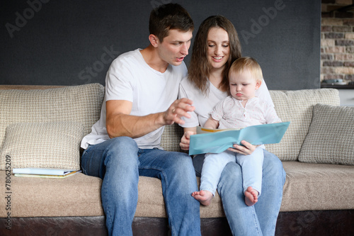 Family: mom, dad sitting on sofa in living room, reading book to baby, showing educational pictures. Young parents spend time with their baby, learning and communicating. Happy family concept,weekend