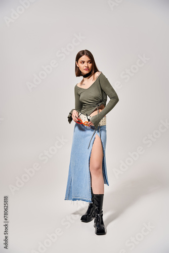 A young woman with brunette hair poses stylishly in a skirt and boots in a studio setting.