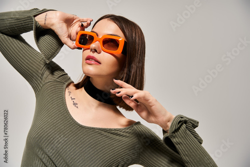 A young woman with brunette hair wearing a green shirt and orange sunglasses poses in a studio setting.