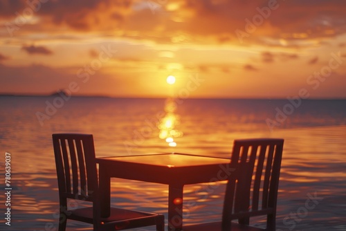 Two chairs and a table positioned on the sandy beach with the sun setting in the background, creating warm reflections on the calm water