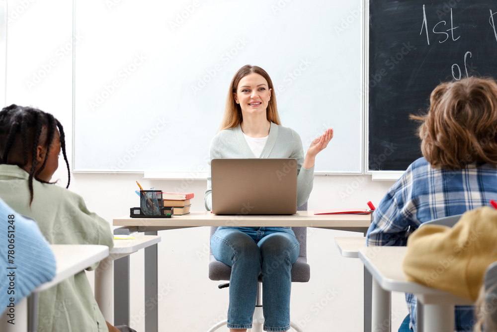 Smiling teacher talking to pupils while sitting at the desk in classroom, using laptop Smiling teacher talking to pupils while sitting at the desk in classroom, using laptop