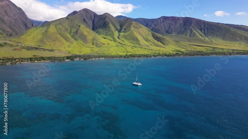 Aerial shot off catamaran boat anchored. Drone pushes forward while gimbals up to reveal dramatic landscape of Maui's northern mountain shores, Hawaii.