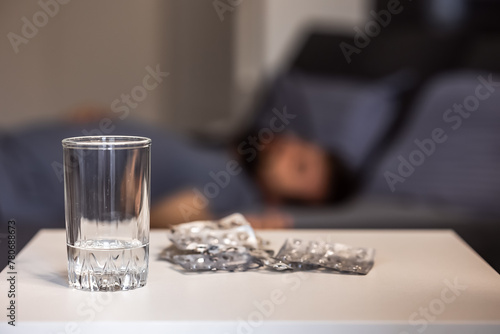 An empty glass and many packs of used empty pills on a table in the bedroom, blurred background. Man lying on bed unconscious or asleep, insomnia, prem pills, used blisters, drug overuse, drug abuse photo