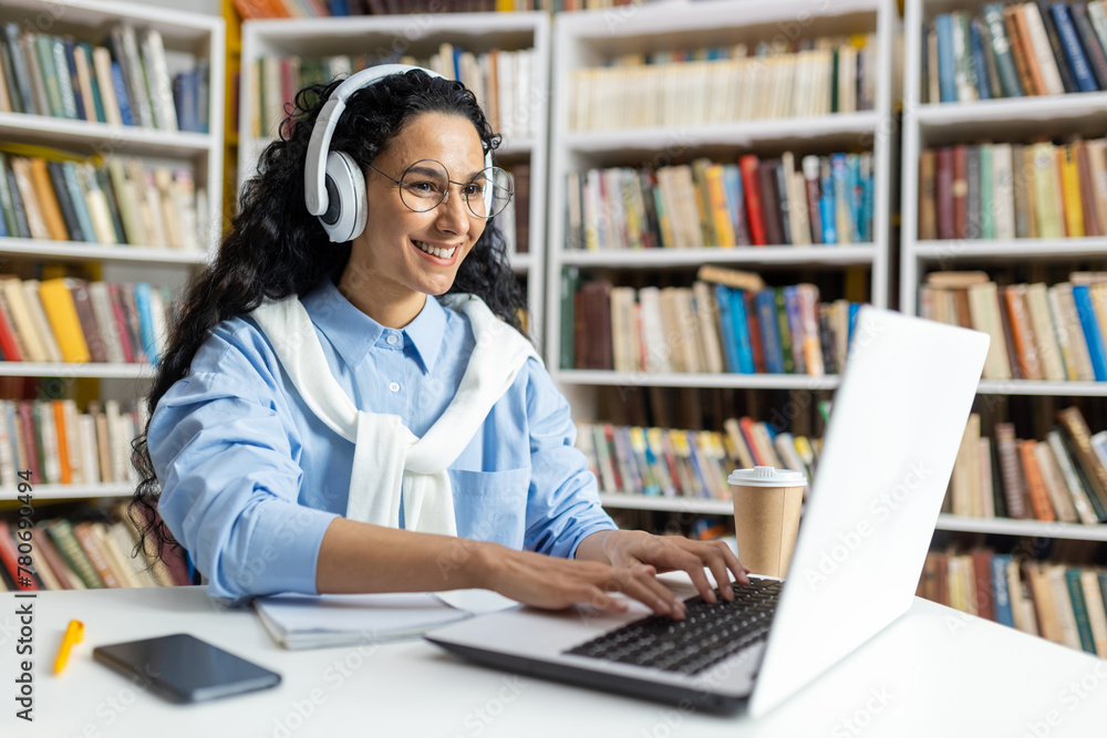 Smiling woman with headphones studying at a laptop in a library setting, surrounded by books and a coffee cup. Smiling woman with headphones studying at a laptop in a library setting, surrounded by books and a coffee cup.