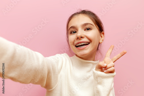 cheerful teenage girl with braces taking selfie and showing peace sign on pink isolated background, happy child taking photo of herself and showing two fingers