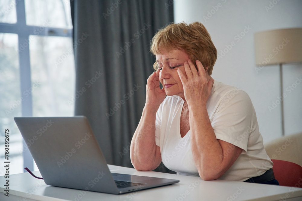 Headache, sitting by laptop. Senior elderly woman is at home in the living room Headache, sitting by laptop. Senior elderly woman is at home in the living room