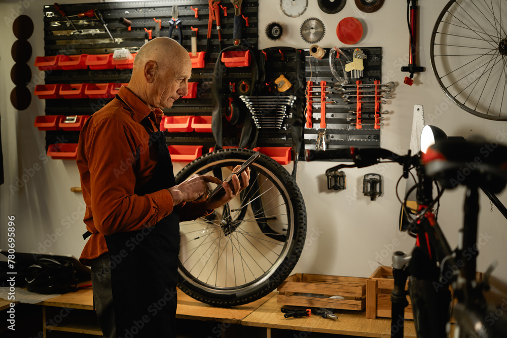 Old male worker holding and repairing bicycle wheel while standing in bicycle workshop or authentic garage