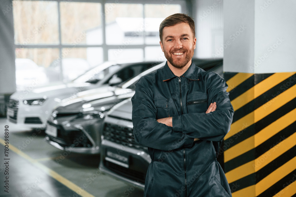 Wiping hands. Repairman in uniform is standing in the garage with cars Wiping hands. Repairman in uniform is standing in the garage with cars