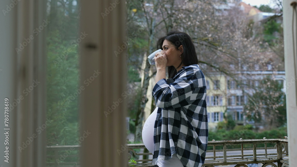 Contemplative pregnant woman stands by apartment balcony sipping tea looking at view from residence, enjoying quiet moment during third trimester