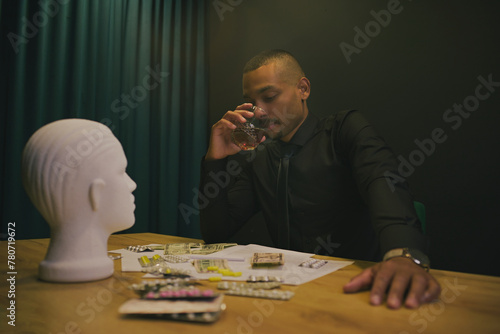 African-American man drinking alcohol whiskey in a casino. There's money scattered on the table, pills mannequin head.