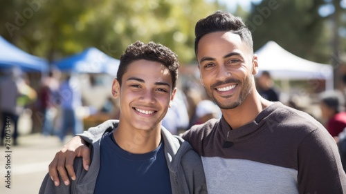A smiling young man with his arm around a younger boy, both happy and outdoors.