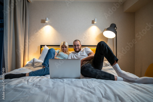 Couple lies on bed, sharing moment of connection over laptop screen, in cozy bedroom setting. 