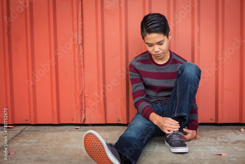 Lonely, sad Asian boy sitting back against a metal wall while gazing down.